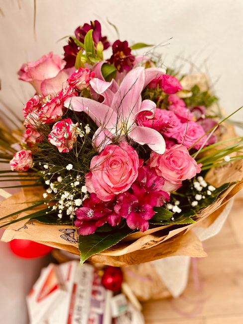 A vibrant floral bouquet featuring pink roses, dark red alstroemeria, and pink gladioli, accented with delicate white baby's breath and lush green foliage. The flowers are arranged in a lively, loose 