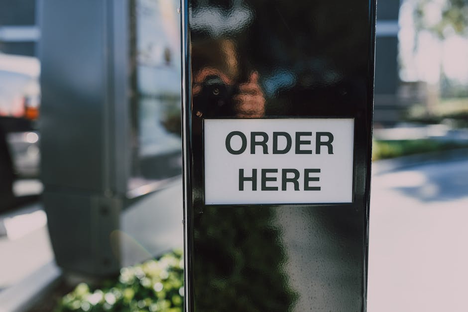 A black outdoor kiosk with a white sign displaying 'ORDER HERE' in bold black letters, situated in a parking area with cars and greenery visible in the background. The kiosk appears to be a drive-thru