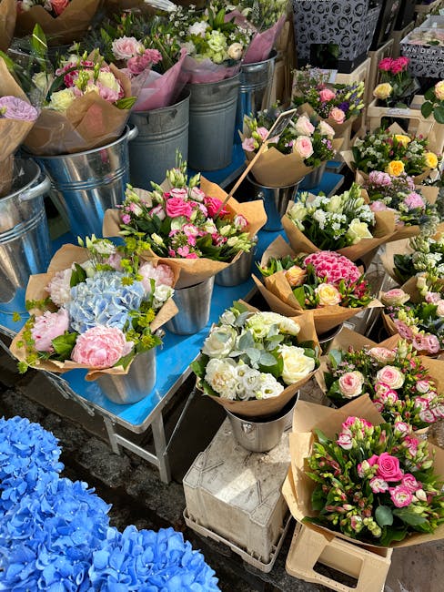 A display of fresh floral arrangements featuring pink, white, and pastel-colored flowers including roses, hydrangeas, and carnations, arranged in small metal buckets lined with pink and brown paper wr