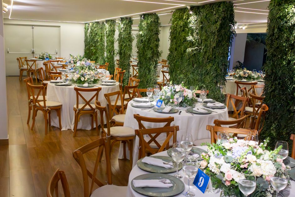 Elegant indoor wedding reception room with round tables draped in white tablecloths, each decorated with lush floral centerpieces featuring pastel pink, white, and lavender flowers, complemented by gr