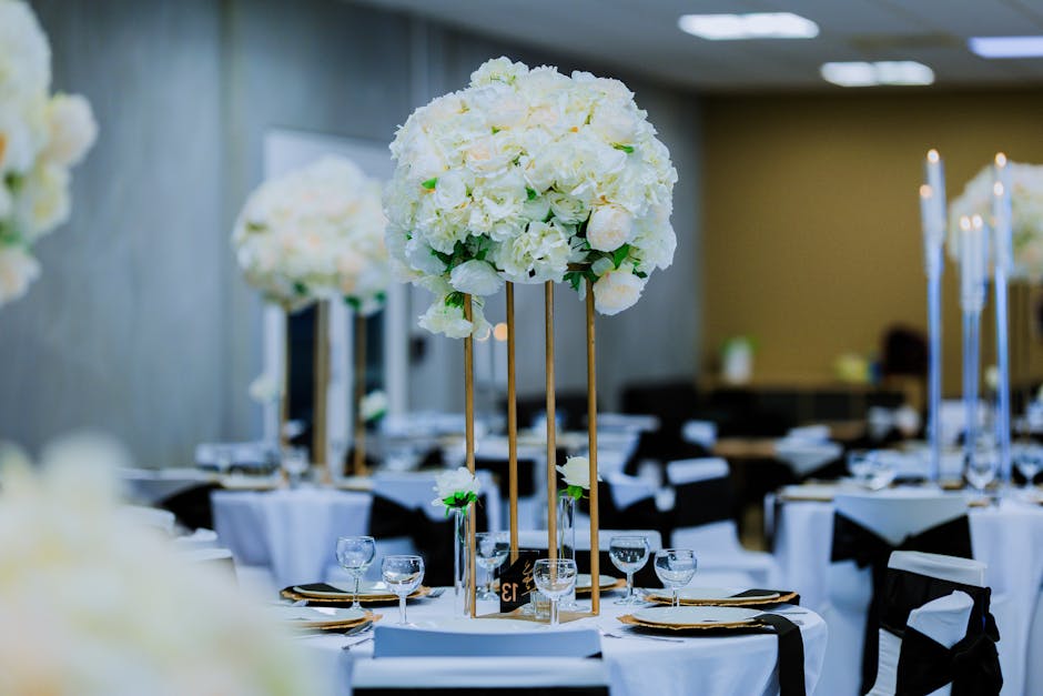 A floral arrangement featuring a large, spherical cluster of white flowers, including roses and hydrangeas, positioned atop tall gold stands in a decorated event space. The arrangement showcases fresh