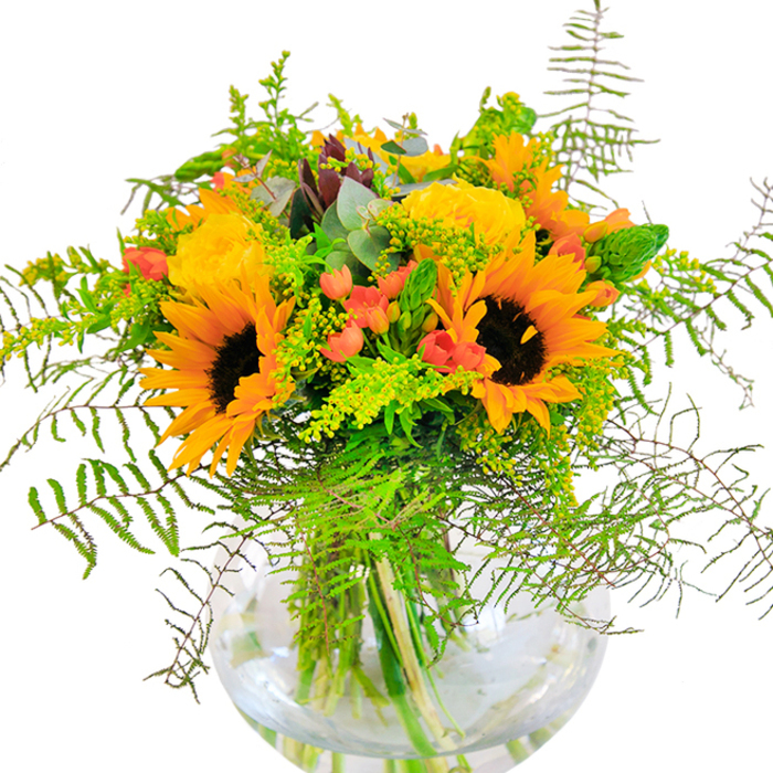 First impressions of this expertly designed bouquet from Harefield are pure sunshine-vivid yellow sunflowers and garden roses nestled together, their luminous petals beaming outward like rays lighting up St Mary's Church green. The sunflowers' bold faces, with textured dark centers and golden fringes, instantly evoke the feel of a perfect summer's afternoon. Around them, lush yellow roses unfurl in tightly-layered spirals, their velvety surfaces catching the light and adding graceful elegance. Deep green foliage weaves throughout-delicate fern fronds gently cascade, their airy structure softening the ensemble and conjuring memories of Harefield's peaceful parkland. Flecks of scarlet hypericum berries punctuate the design, their glossy skins glinting like treasures among the petals and greens, creating a striking contrast. Bright yellow solidago filler and long, slender green floral spikes contribute both volume and natural flow to the composition, while cool eucalyptus leaves introduce a touch of blue-green, refreshing the scene. Every stem aligns neatly in a round, clear glass vase, its transparency revealing a tangle of green stems submerged in shimmering water. The bouquet's scent is a subtle blend of crisp greens and gentle florals, reviving the senses and evoking sun-warmed fields. Perfect for birthdays, anniversaries, or gatherings in Harefield, this arrangement is a vivid tribute to happiness and togetherness-a handcrafted piece designed to bring joy and a touch of natural luxury into the heart of local homes.
