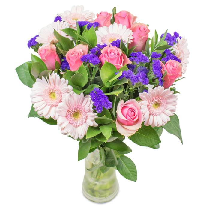 An exuberant, expertly arranged bouquet sits elegantly within a transparent glass vase, perfectly positioned against a crisp white backdrop that accentuates every shade and detail. At the heart are 7-9 medium pink roses, their petals unfurling in tight, spiral formations-some buds barely open, others just at the peak of bloom-demonstrating pristine classic form and romance. Gently framing them are 5-7 pale pink gerbera daisies, each displaying large, round faces and layers of slender petals, with creamy centres hinting at lightness and serenity. Interspersed throughout, rich violet clusters of statice provide striking texture and a vibrant pop, their papery blooms reminiscent of flower meadows on the edges of Harefield's green expanses. Beneath and between these star flowers rests an opulent bed of deep green foliage-each glossy leaf enhancing the bouquet's sense of life and freshness, as if recently gathered from nearby woodlands. The clear vase reveals clean-cut stems and shimmeringly fresh water, emphasising a just-delivered, celebratory quality. This arrangement, suitable for birthdays, anniversaries, or heartfelt Mother's Day gift-giving around Harefield, effortlessly conveys joy, warmth, and a personal touch steeped in local expertise.