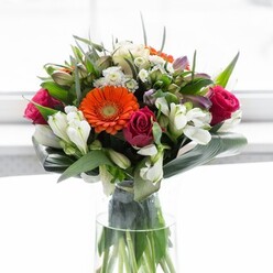 This beautifully crafted arrangement by Harefield Florist channels both artistry and local warmth, seamlessly blending creamy peach and fiery orange gerberas with sumptuous magenta roses. Each bloom captures the gentle light pouring in through a bright, soft background evocative of a tranquil morning in Harefield, perhaps inspired by vistas over the village green or nearby woodlands. The expansive, dark green aspidistra leaves are folded in careful arcs, gracefully enveloping the flowers and echoing the lush hedges found along Harefield's lanes. Between the central blooms, wisps of delicate white alstroemeria and tiny spray roses intermingle with subtle hints of blush pink, filling the bouquet with vibrancy and texture. The clear glass vase, tall and elegant, exposes the fresh green stems, reinforcing a sense of organic vitality and local, farm-fresh appeal. Gentle sprigs of fine greenery thread between petals, adding movement and volume, while the interplay of colour and texture suggests thoughtful arrangement from an experienced florist steeped in community knowledge. The bouquet radiates optimism-a perfect centrepiece for a brunch, a symbol of fresh beginnings, or a uniquely Harefield gesture of care and appreciation delivered throughout the area. Its fragrance, a subtle blend of rose and lily with cool greens beneath, invites memories of country air and new seasons.